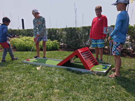 Four kids taking turns at a numbered mini golf hole beside the water