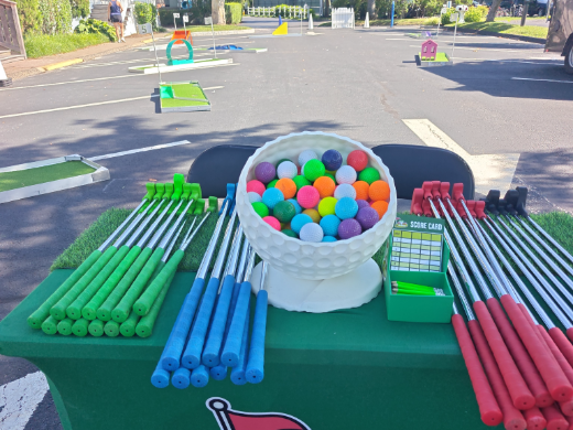 Putters, colored golf balls and scorecards laid out on the Mini Golf on the Go equipment table, course set up behind