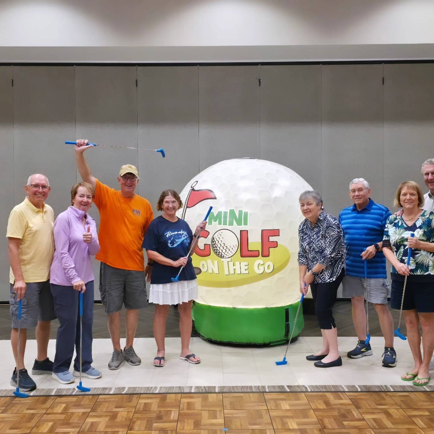 A group posing with putters beside the giant Mini Golf on the Go ball in a ballroom