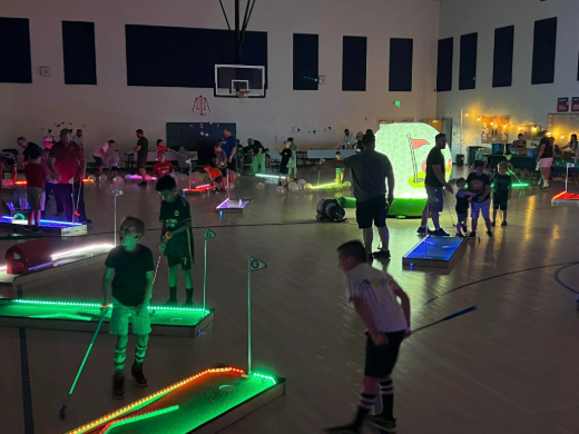 Glow-in-the-dark mini golf night in a school gym, every hole outlined in LED light with the lit golf ball behind