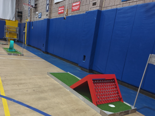 Portable mini golf hole with a red obstacle set up indoors on a school gym floor
