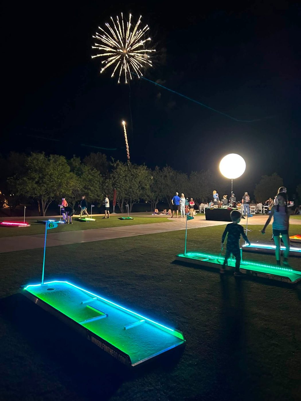 LED-lit mini golf holes glowing green and blue under fireworks at an evening event