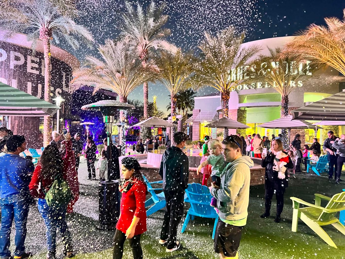 Families standing in falling snow at an outdoor shopping plaza holiday night