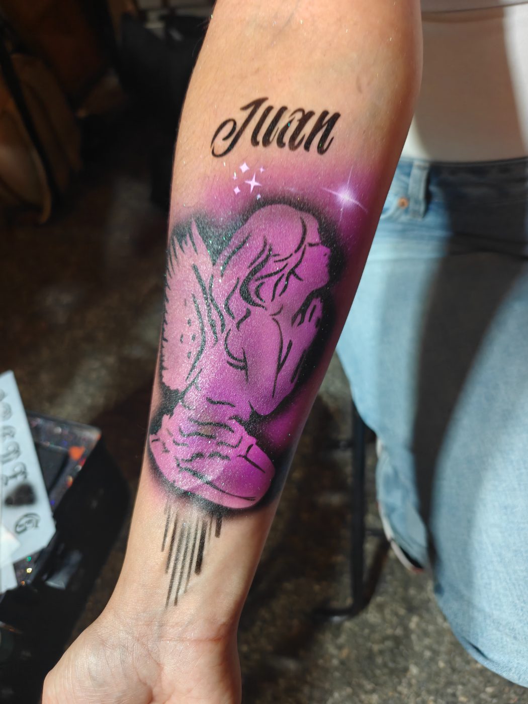 Airbrushed angel in purple on a forearm with the name Juan lettered above it